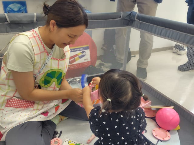 mama's marche（相浦コミュニティセンター）の託児ブースで、スタッフが笑顔で子どもと一緒におもちゃで遊んでいる様子